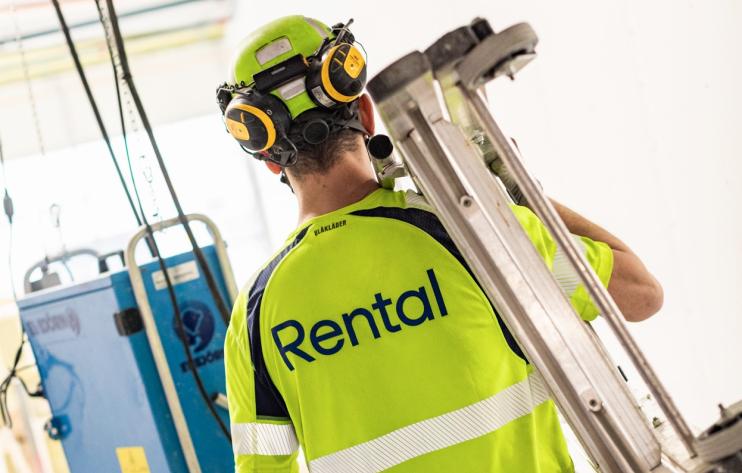 En byggarbetare sedd bakifrån i gul varselväst med texten ”Rental” och hjälm, bärandes på ett stege över axeln på en byggarbetsplats.